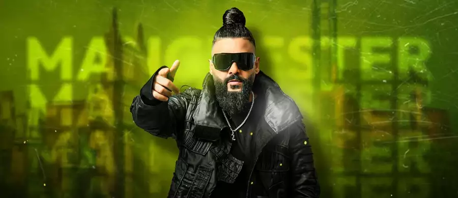Sasy with his hair in a bun wearing dark sunglasses. The background is green with the word 'Manchester' displayed three times.