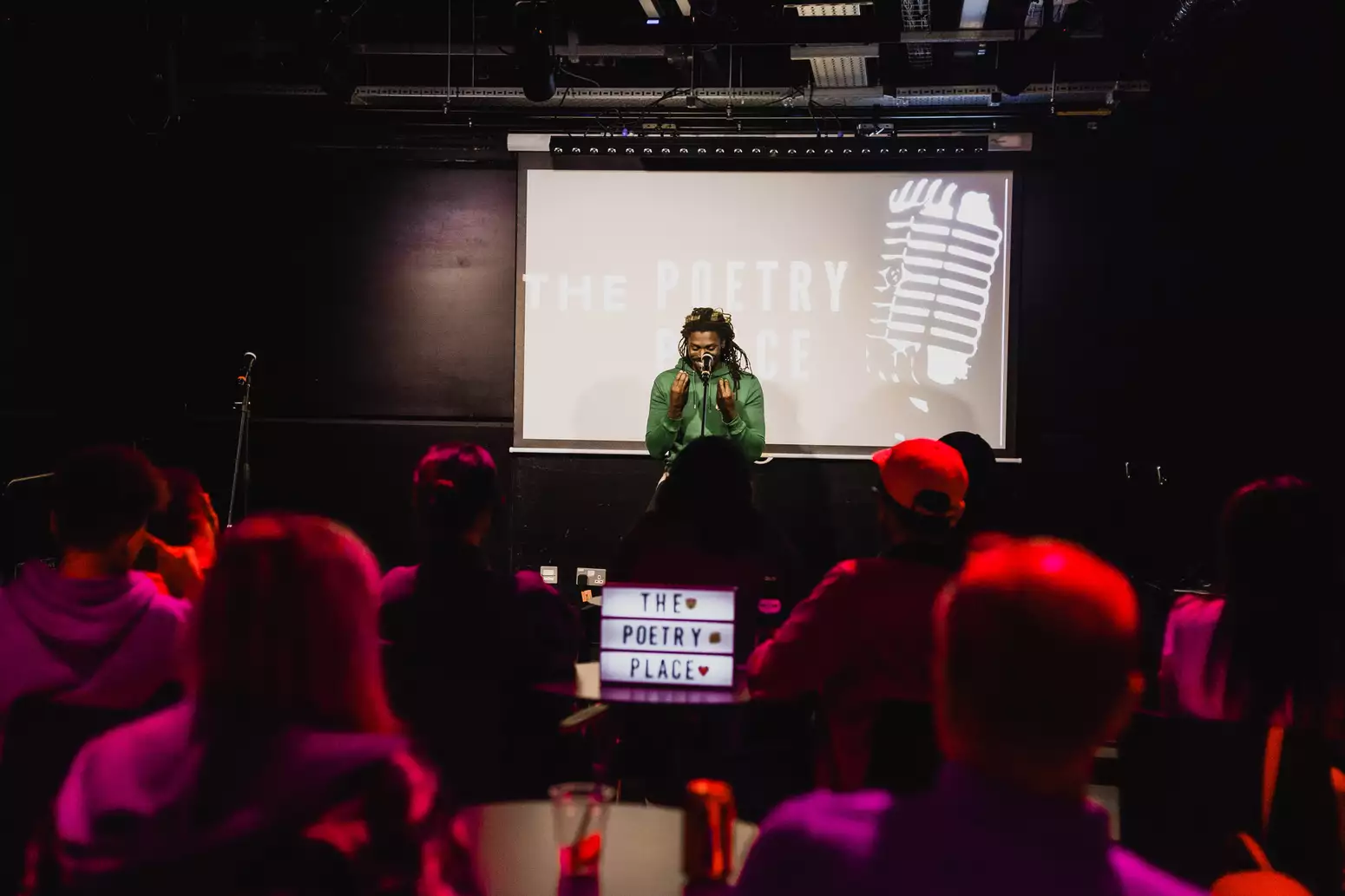 A poet performs on stage at a spoken word event as an audience watches in a dimly lit venue. There is a screen behind them which says 'poetry place'.