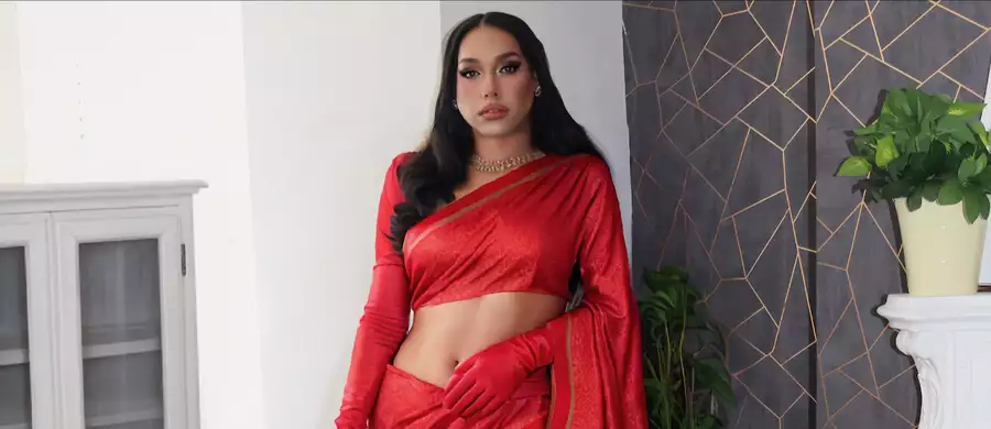 Lucky Roy Singh stands confidently indoors wearing a striking red saree with gold accents and matching red gloves. Their long, wavy black hair is styled neatly, and they accessorise with gold earrings and a chunky necklace. The backdrop features a geometric-patterned dark wall, a white cabinet, and potted plants, creating a blend of traditional elegance and modern decor.