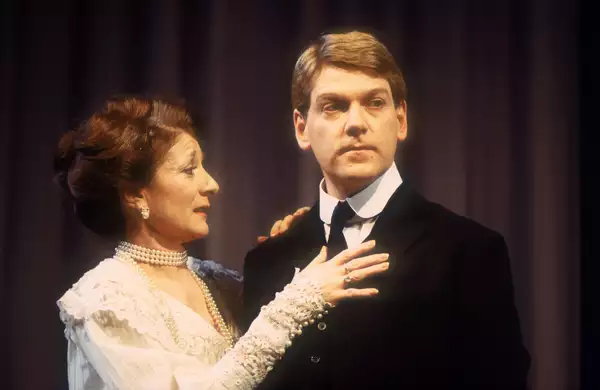 Jane Lapotaire and Kenneth Branagh on stage as Gertrude and Hamlet. Jane is wearing a white lace dress with her hands on Kenneth's chest. He is wearing a suit and looking off-camera.