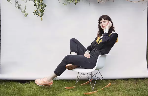 Anna Meredith sits in a modern white rocking chair outdoors, posing in front of a white backdrop with green branches hanging above. She wears a black shirt with yellow detailing, dark jeans, and peach sneakers.