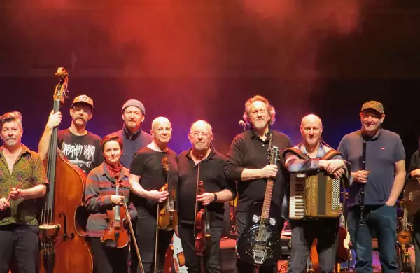 A group of musicians stand together on stage holding their instruments, including fiddles, guitar, accordion, flute, upright bass, and banjo. Warm red stage lighting creates a lively atmosphere behind them.