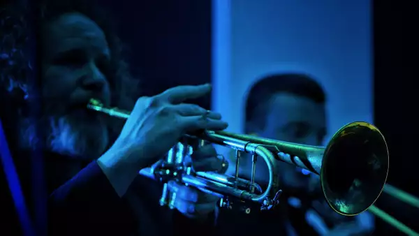 Close-up photo of a person playing a trumpet. The lighting is blue.