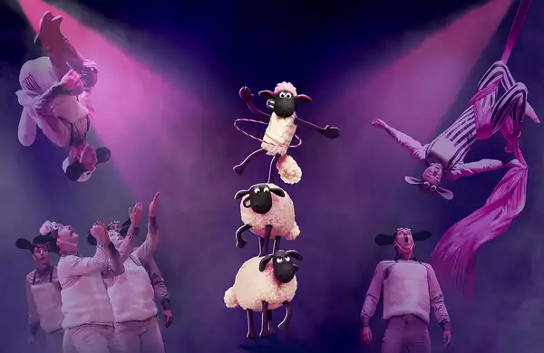 Performers in sheep costumes execute acrobatic stunts and aerial silk acts under dramatic pink lighting. In the center, three animated sheep from "Shaun the Sheep" balance in a tower, with the top one hula-hooping.