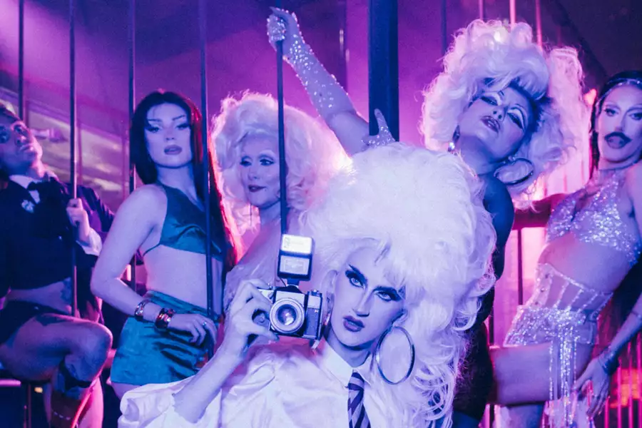 A vibrant group of drag performers pose dramatically under purple and pink stage lighting. The central figure, with voluminous white hair and bold makeup, wears a white shirt, black corset, and fishnet tights while holding a vintage camera. Surrounding them are five other performers in glamorous, theatrical outfits, striking expressive poses behind a metal cage-like structure on stage. The scene radiates energy, glamour, and queer cabaret flair.