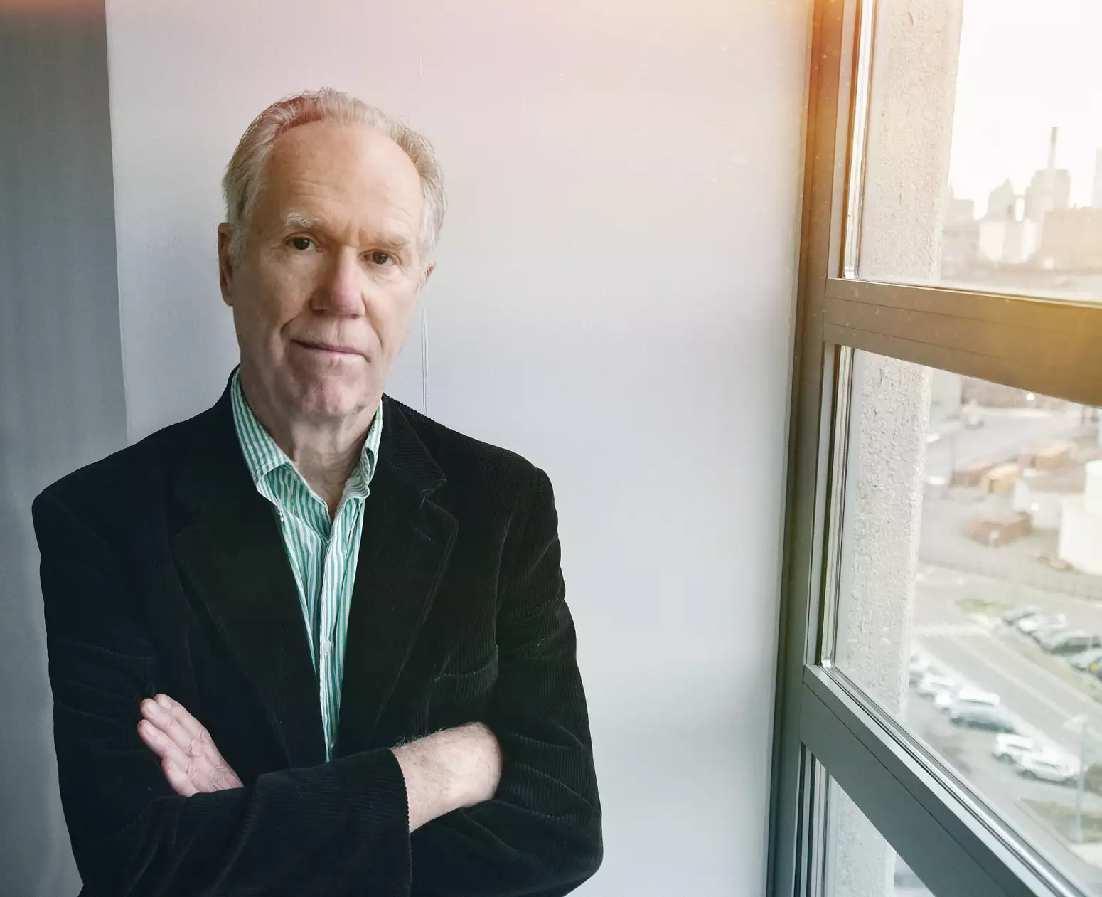 Loudon Wainwright III stood with his arms folded by a window. He is wearing a black velvet suit jacket.