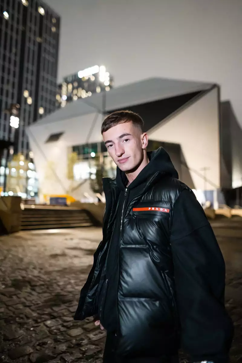 Close-up photo of Azyr wearing a puffer jacket stood outside Aviva Studios at night. The background is blurry.
