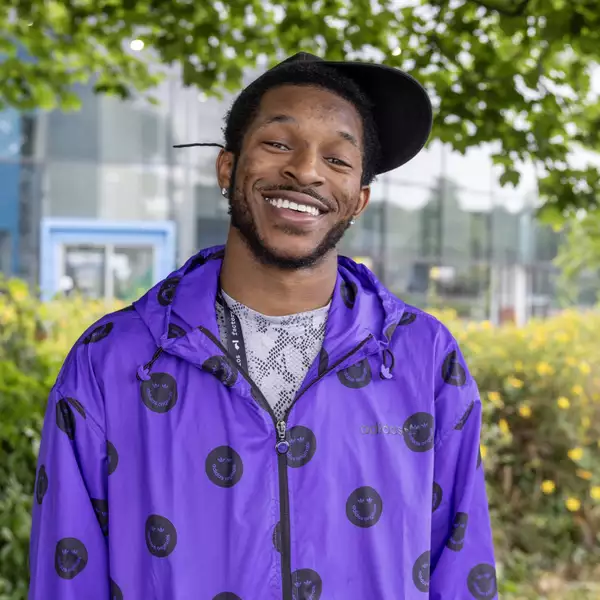 headshot of Zeriah Otutu wearing a black cap and a bright purple waterproof with black smiley faces on it. They are stood outside under a tree.