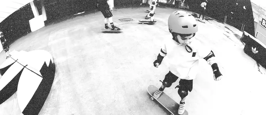 Black and white image of a kid learning to skateboard on the Adidas skateboard ramp, wearing a helmet