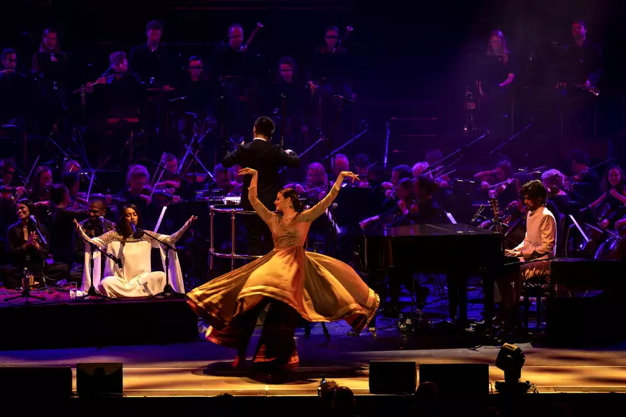 Orchestral performance featuring a qawwali singer, pianist, conductor, dancer in flowing dress, and a full orchestra in the background.