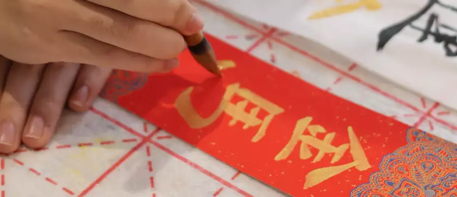 Hand writing Chinese calligraphy in gold ink on a red paper.