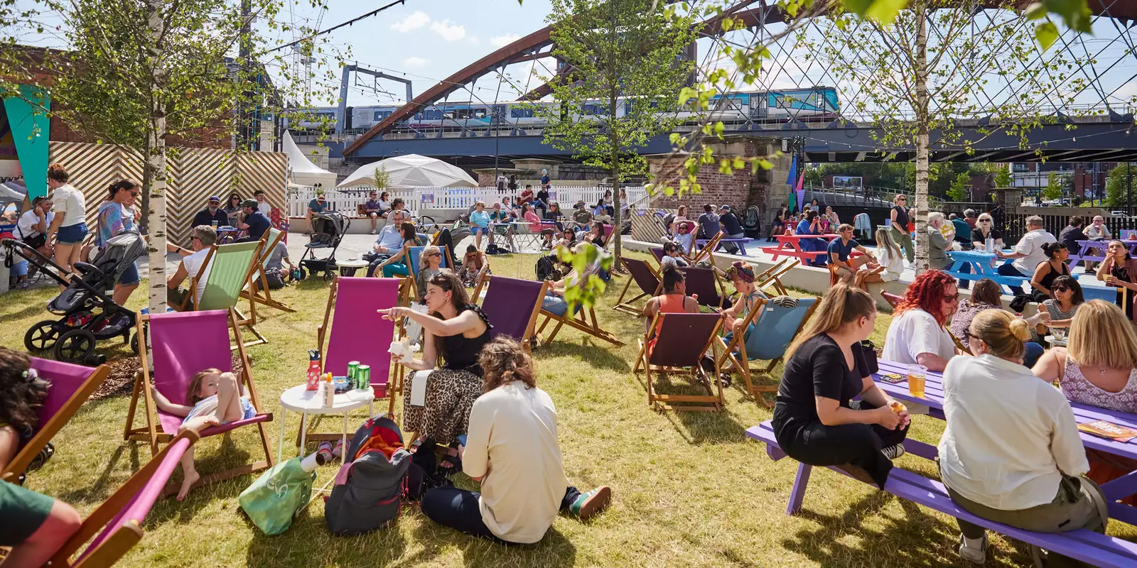 Factory Square filled with people sat on the grass and deckchairs, drinking and chatting. The sun is shining.