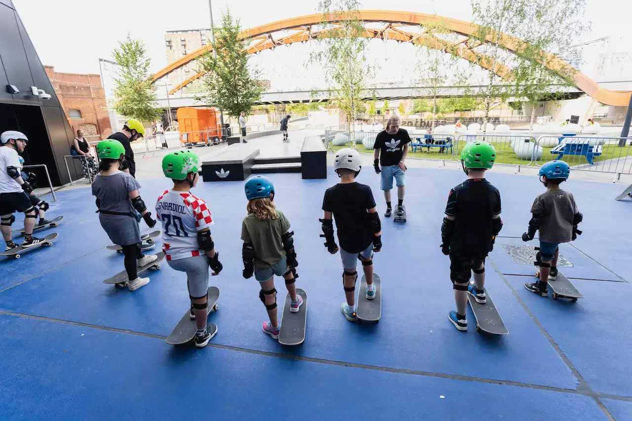 A group of children with skateboards and helmets, taking part in a skateboarding class outside Aviva Studios as part of Build Manchester