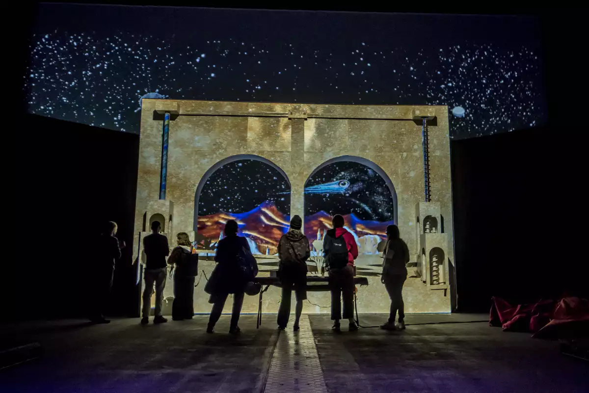 Studio Tonto's installation in the South Warehouse – showing a group of people standing in silhouette, facing a brightly lit archway featuring colourful patterns, and a cosmic background featuring stars and a large bird.