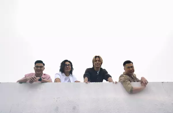 Four members of Rubberband leaning over a grey wall outside, smiling. The background is overcast.