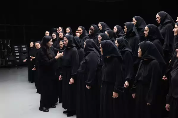 A photo of artist Marina Abramovic talking to a group of performers dressed all in black as if at a funeral