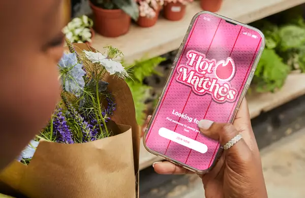 A person holding a bouquet of flowers is using a smartphone displaying the "Hot Matches" dating app. The app screen features a bright pink background with the tagline “Looking for love?” and a button labeled “Join Now.”