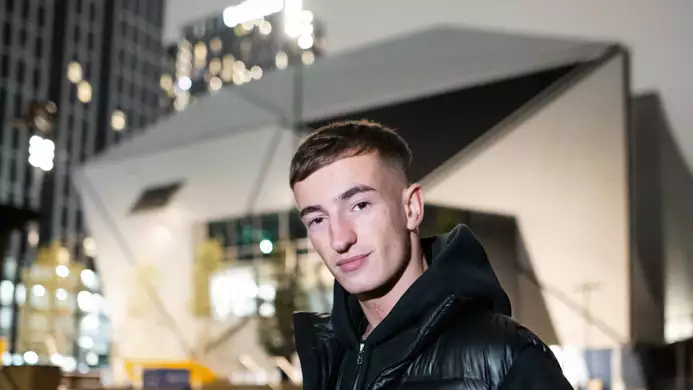Close-up photo of Azyr wearing a puffer jacket stood outside Aviva Studios at night. The background is blurry.