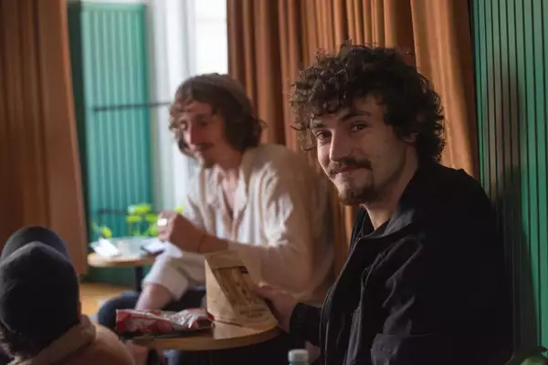 Three young men are sat eating sandwiches with a curtain behind them. One is looking at the camera while the other two are out of focus.