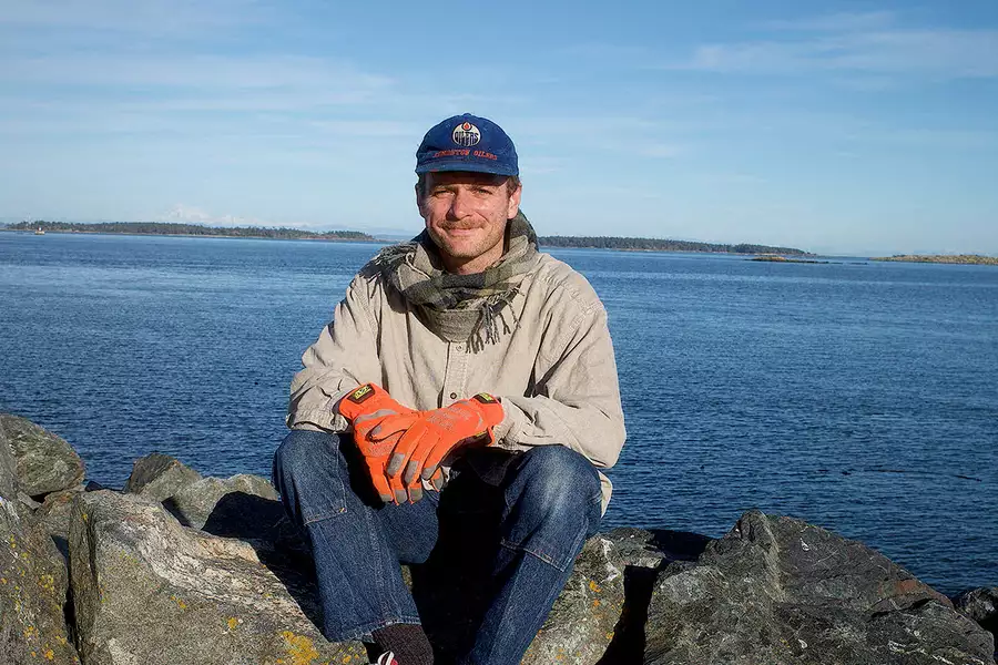 Mac DeMarco sitting by the sea on some rocks, wearing a cap and big orange gloves