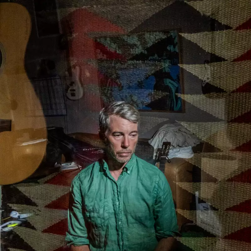 Bill Callahan wearing a green shirt sitting indoors, overlaid with a double exposure of a guitar and patterned rug.