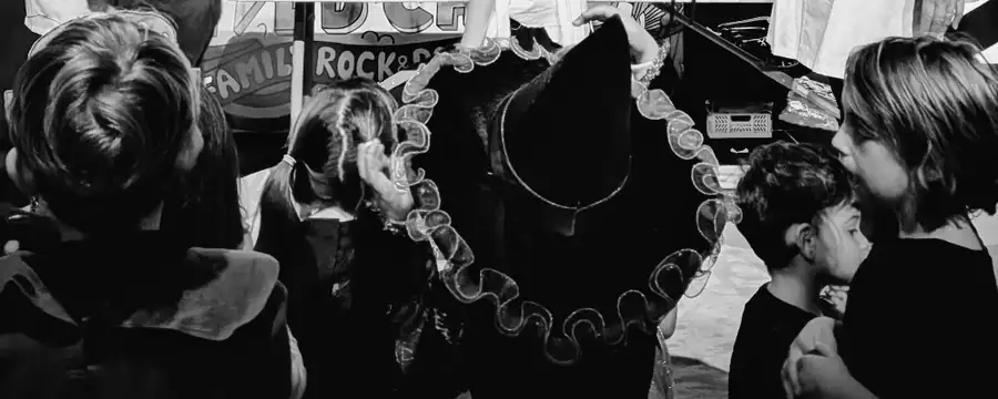 A black and white photo of children watching a performance. One is wearing a witch fancy dress hat.