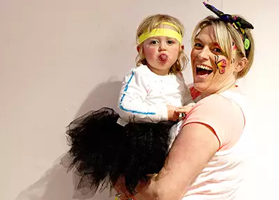 A child pulling out their tongue wearing a black ballerina tutu – who is being held by a parent wearing face-paint