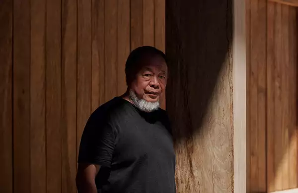 Ai Weiwei wearing a black t-shirt, stood in front of a wooden backdrop. There is a shadow across his face.