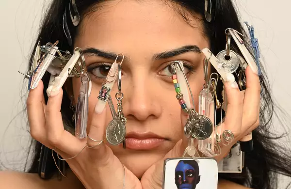 A person with decorated nails holding their face, featuring various keychains and trinkets attached to their nails.