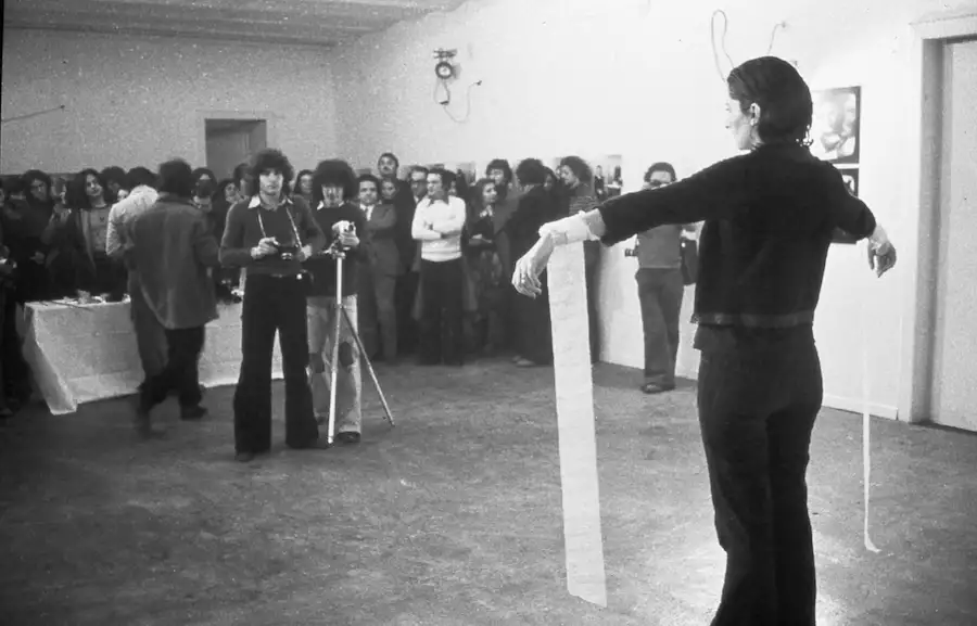 Black and white photograph of Marina Abramović performing Rhythm 0. She is standing with her arms outstretched in front of a crowd of people.