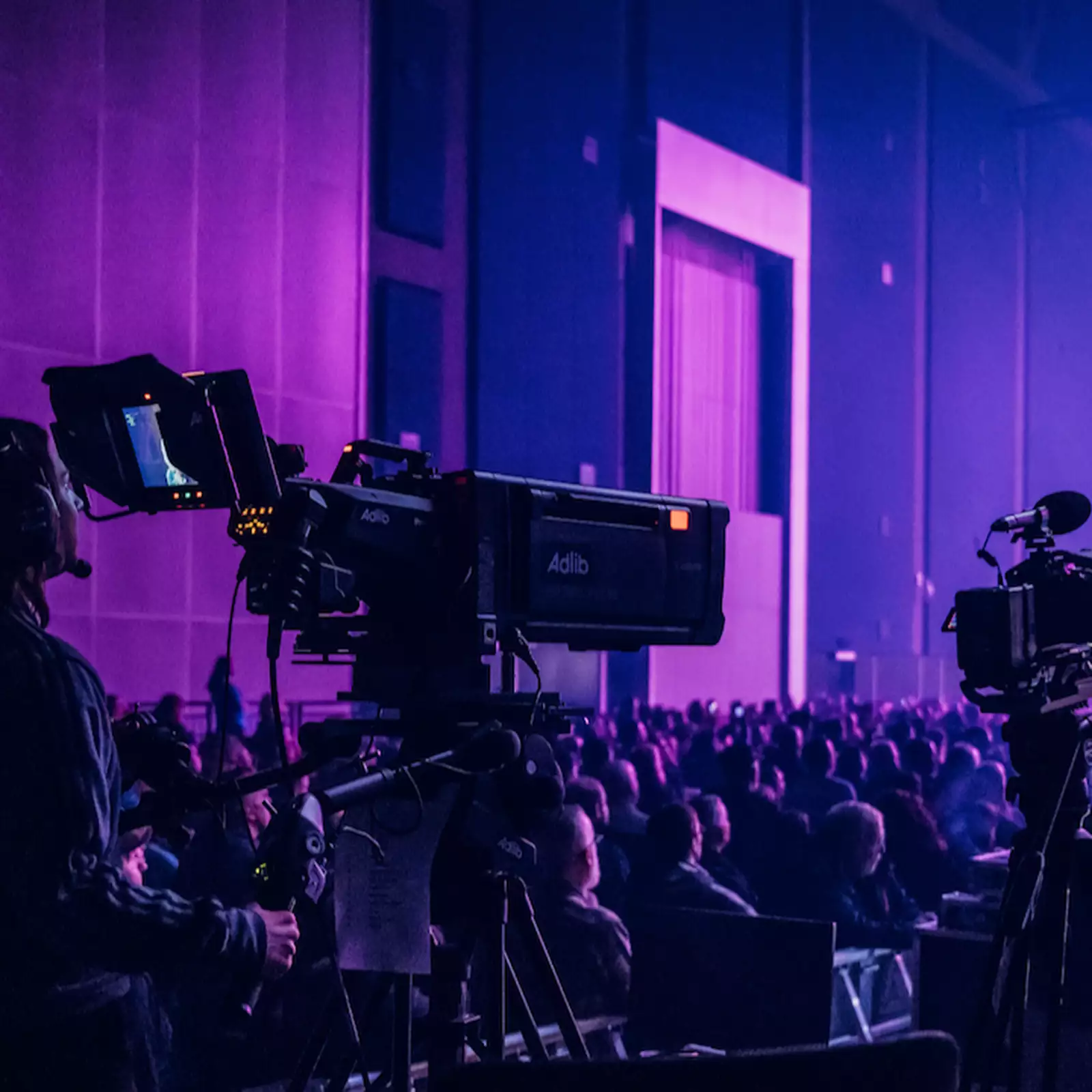 A camera operator films a live event in the Warehouse, which is filled with people. There is dramatic purple lighting with multiple cameras and bright stage beams.