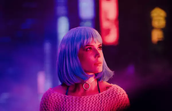 Halsey with a short, straight, light blue wig and wearing a silver ring choker looks upward under neon purple and pink lights in a futuristic, cyberpunk-style setting.