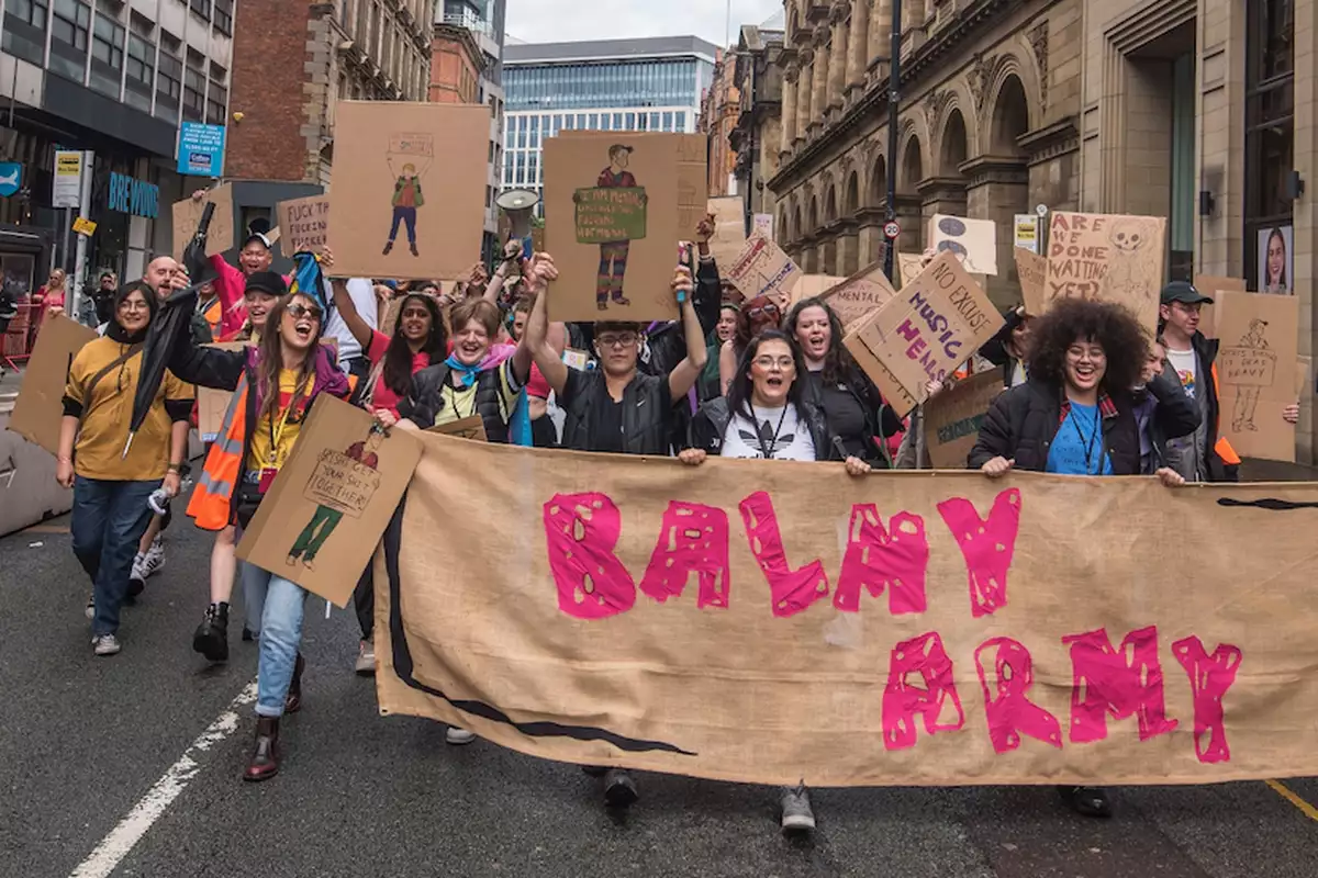 A group of young people marching through Manchester City Centre with a huge banner that reads 'Balmy Army'. Many are holding placards and megaphones.