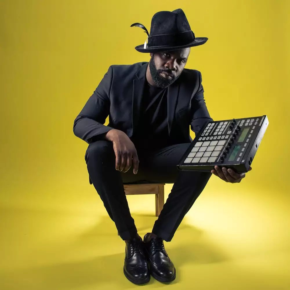 Photo of Michael Asante wearing a black suit and hat, sat on a small wooden stool holding a mixer. The background is bright yellow.
