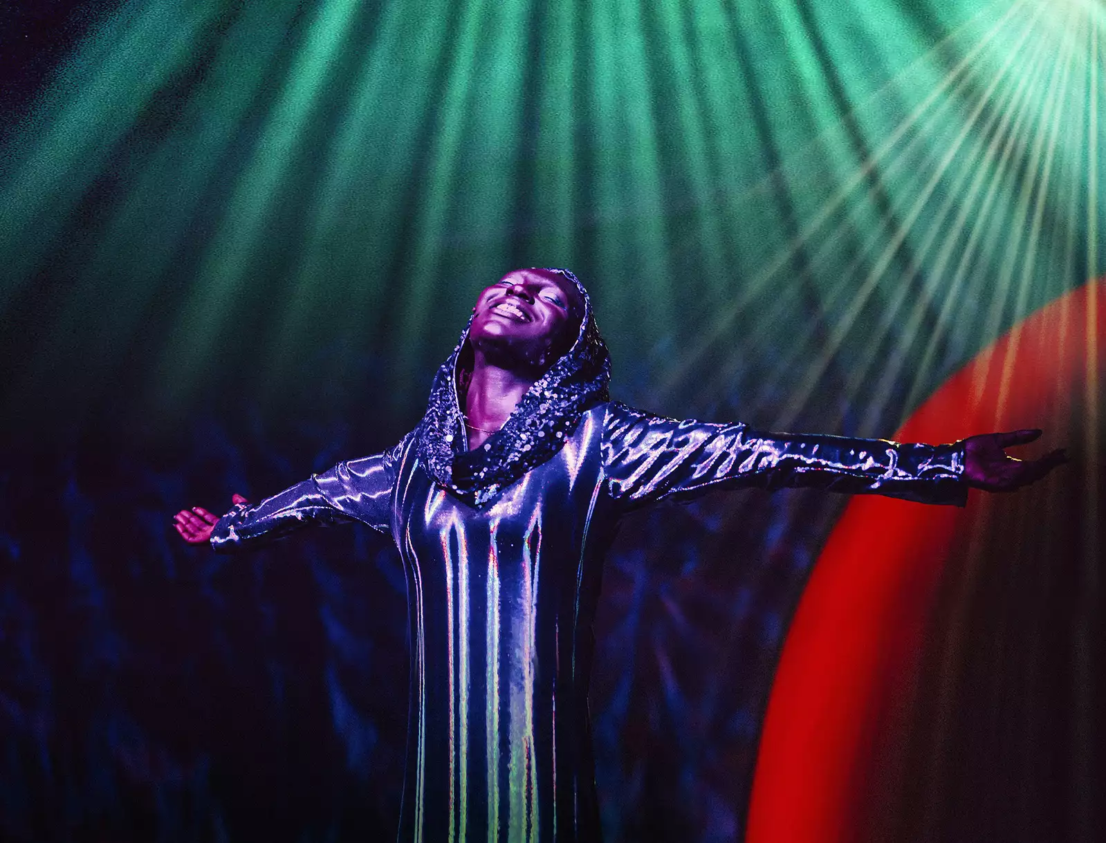 A performer smiles with their arms outstretched and eyes closed during WAKE. They are wearing a silver, glittery dress and head covering.