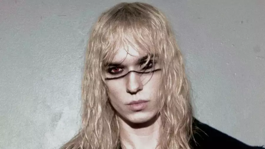 2hollis faces the camera with long blonde hair and bold black eye makeup, wearing a black jacket, striped top and cross necklace against a grey background.