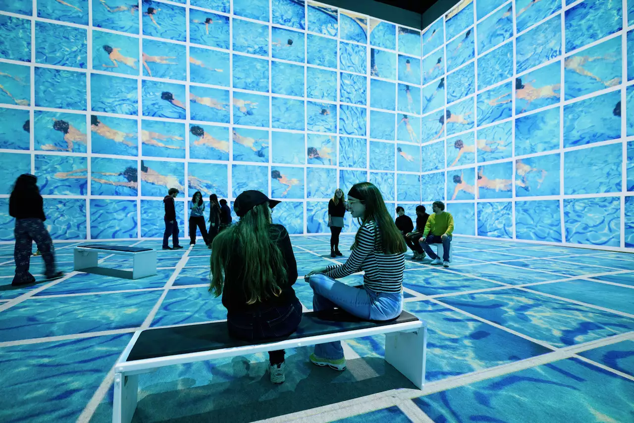 An image from David Hockney: Bigger & Closer (not smaller & further away) showing two people sat on a bench talking, surrounded by his swimming pool artwork