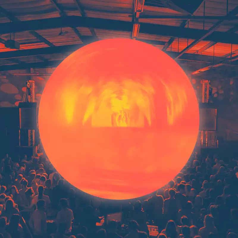 A large, glowing orange orb is suspended above a crowd in a dark indoor venue, casting warm light over the audience and the industrial-style ceiling.