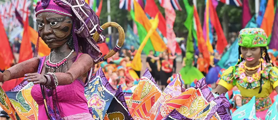 Colourful parade with large puppets, vibrant costumes and waving flags in a crowd.