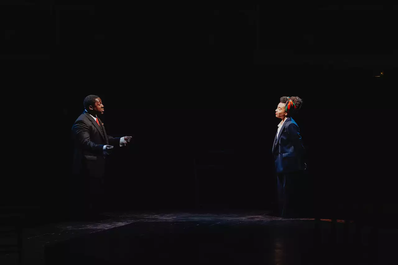Two actors mid-conversation, stood far apart wearing suits on the stage at the Royal Exchange
