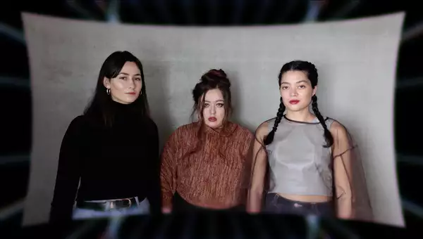 Three members of Keiken staring at the camera, stood in front of a large, grey piece of material.