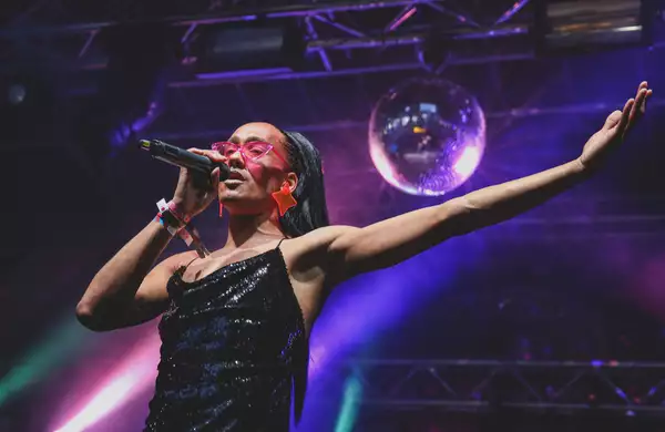 A person singing on stage with a disco ball and purple lighting. They are wearing a black glittery dress and pink sunglasses.