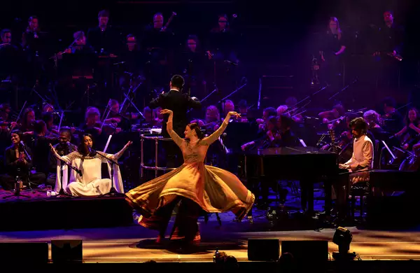 Orchestral performance featuring a qawwali singer, pianist, conductor, dancer in flowing dress, and a full orchestra in the background.