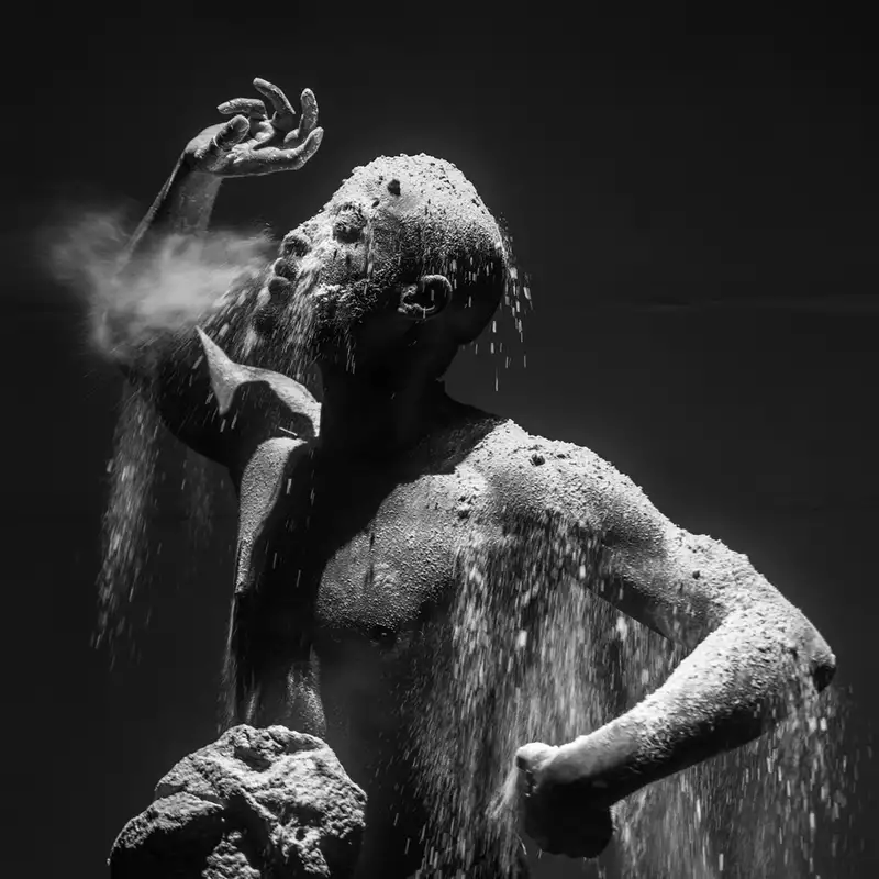 A dramatic black-and-white photograph of a man covered in dust and sand, with particles cascading over his body as he exhales forcefully.
