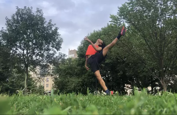 A horizontal photo of ​​Benedict Nguyễn, wearing black shorts, orange tank, and black and orange sneakers, standing on right leg with left leg raised to the side, left arm bent in front of her torso, head tipped back, in a field with green grass and trees, with buildings in distant background.