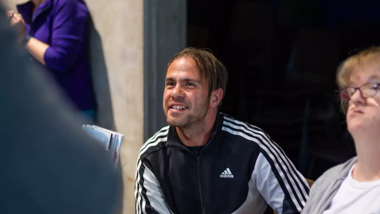 A smiling man wearing a black and white Adidas track jacket is seated indoors, engaging with someone off-camera.