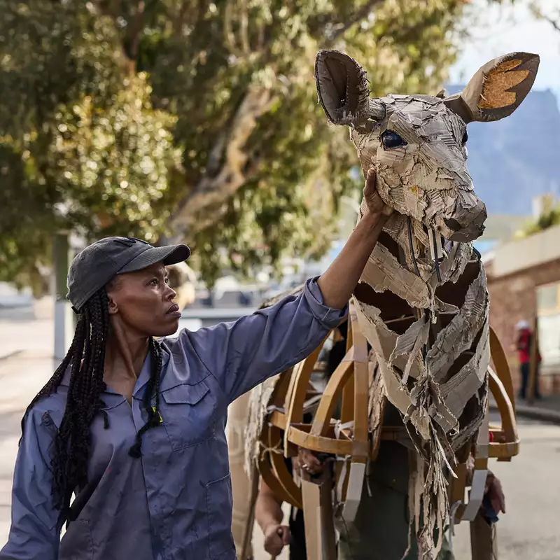 A woman in a cap touches the head of a life-sized giraffe puppet made from cardboard and wood, as part of The Herds. The background shows an urban environment with people watching and a scenic view of trees and mountains.