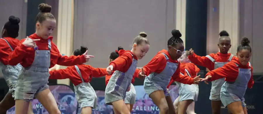 A group of children from Energy Dance mid-dance. They are wearing blue dungarees and orange/red jumpers.