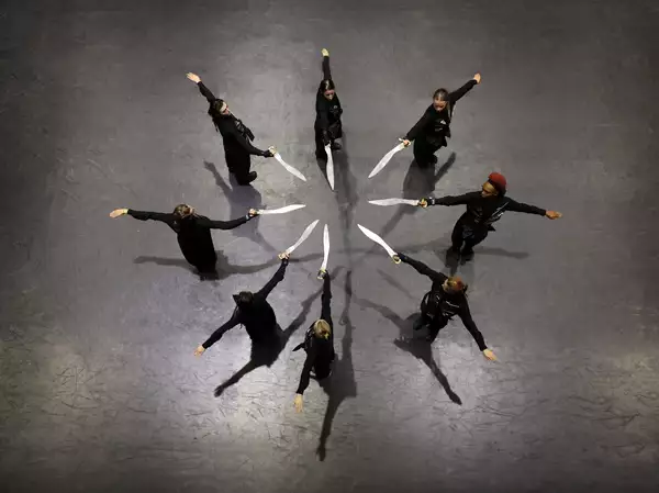 An aerial photo of dancers in a circular formation each pointing knives towards the centre of the group
