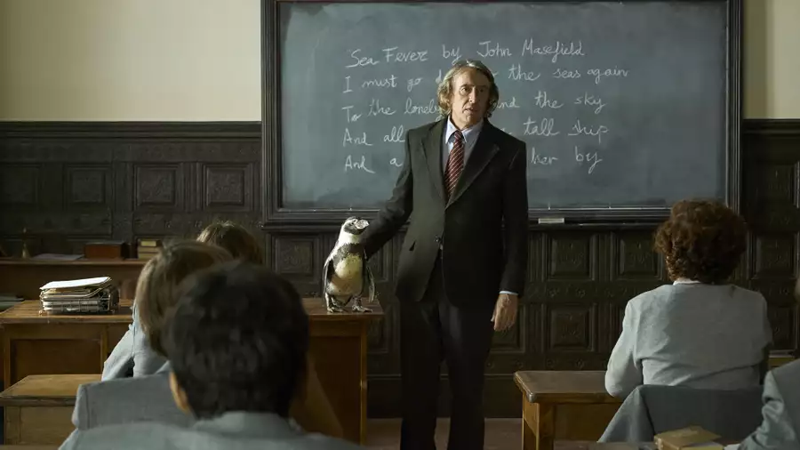 A still from The Penguin Lessons showing a classroom scene with Steve Coogan. He is wearing a suit and standing in front of a blackboard. He rests one hand on a penguin standing on a wooden desk. Students in grey uniforms sit at wooden desks.